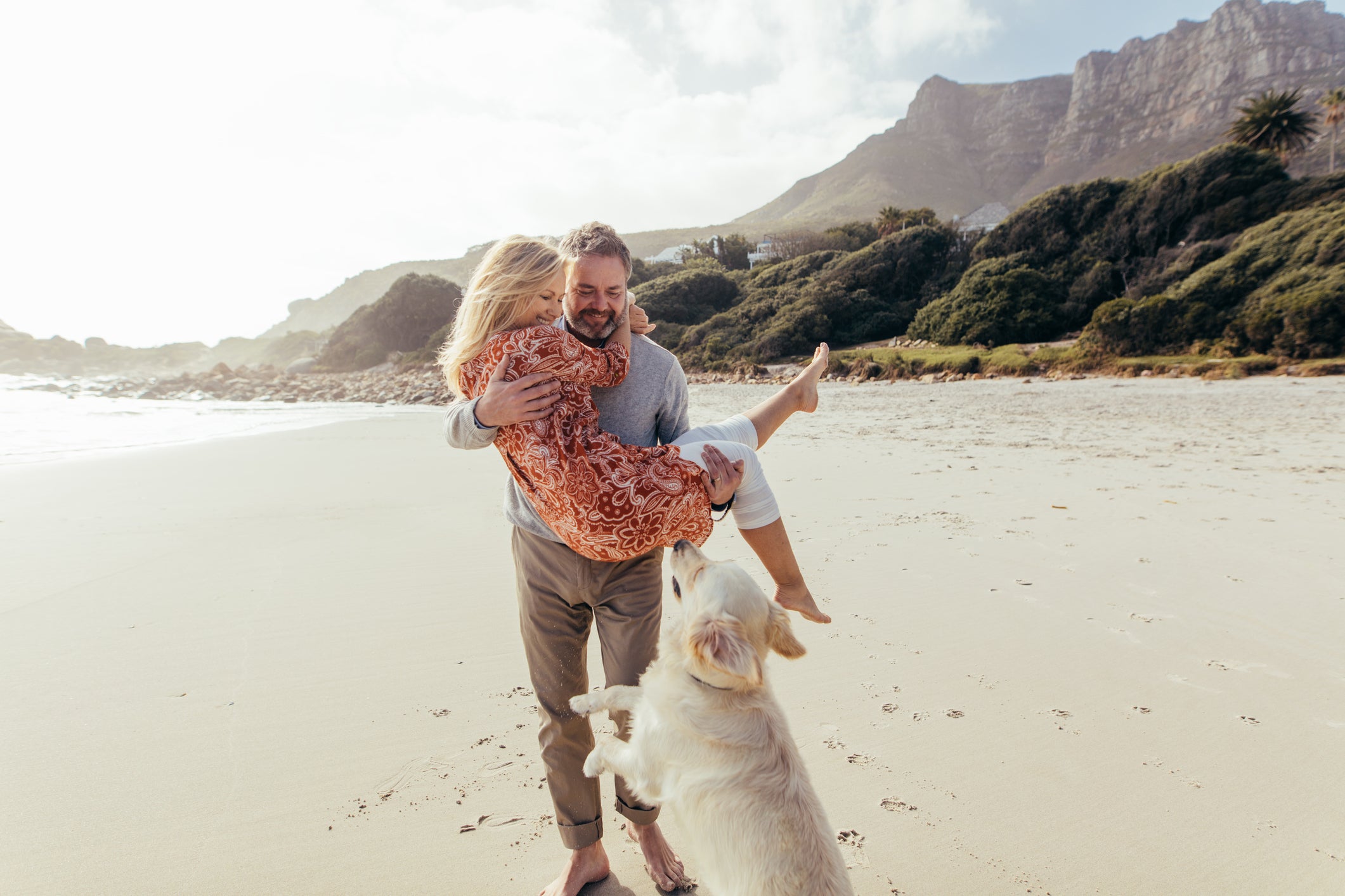 A husband and wife in their elder years at the beach with their dog. The man is carrying his wife while the dog is jumping up.